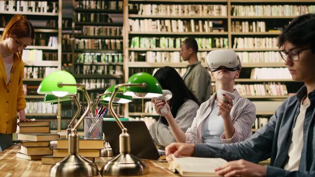 Student wears VR glasses and holds controllers in university library, using immersive technology beside classmates with open books.