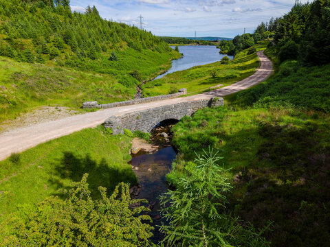 Scenic dirt road winding across old stone bridge in lush green valley with lake