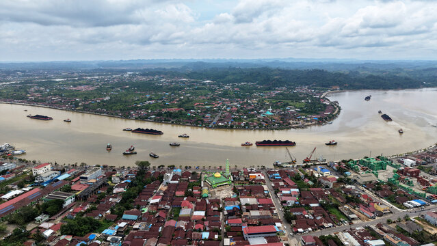 Aerial view of a river town featuring a prominent mosque, coal barges on the river, and dense residential housing, Berau Regency, East Kalimantan, Indonesia.