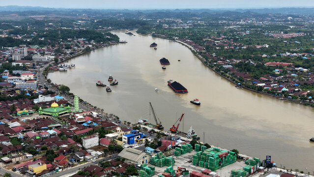Aerial view of coal barges and tugboats navigating the river near the port facilities and residential areas in Berau Regency, East Kalimantan, Indonesia.