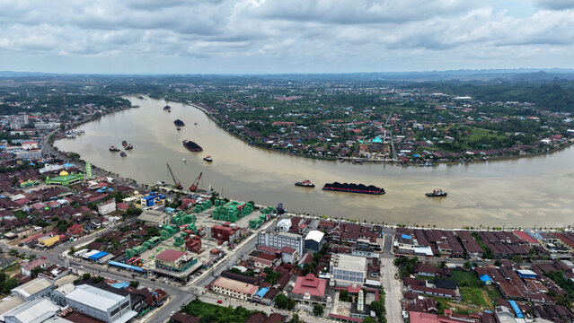 Aerial view of the Segah River with coal barges, industrial port facilities, and residential areas with mosques under a cloudy sky in Berau Regency, East Kalimantan, Indonesia.