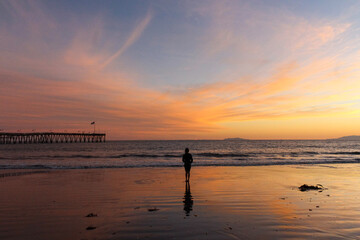 Fototapeta premium Person Standing on Beach at Colorful Sunset Near Ocean Pier
