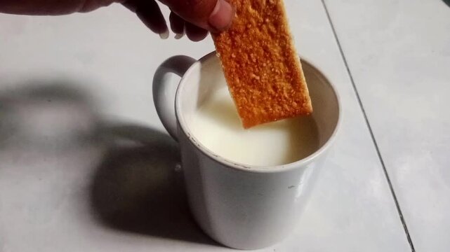 Close-up footage of a hand dipping a biscuit into a cup of milk. Simple breakfast and snack concept with natural movement and food preparation theme.