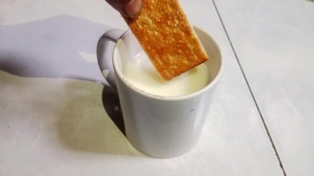 Close-up footage of a hand dipping a biscuit into a cup of milk. Simple breakfast and snack concept with natural movement and food preparation theme.
