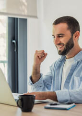 Fototapeta premium Young man working remotely on his laptop, smiling and raising his fist in celebration of achieving a goal or winning an online activity from his modern home office setup