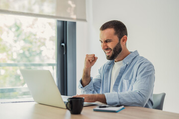 Naklejka premium Young man jubilantly celebrating a win or achievement while working on a laptop at a desk, showing excitement and triumph in his remote workspace