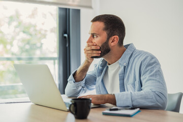 Naklejka premium Man covering his mouth with a hand, sitting at a desk and looking at a laptop screen with a shocked and concerned expression, reacting to a mistake or unexpected negative event