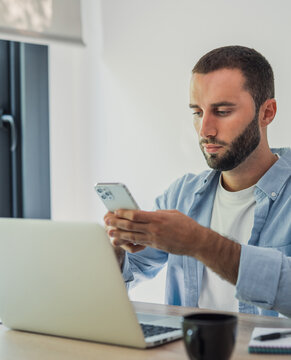 Young man using a smartphone and laptop simultaneously, managing tasks and communicating online from a modern office, blending digital platforms for business and personal productivity