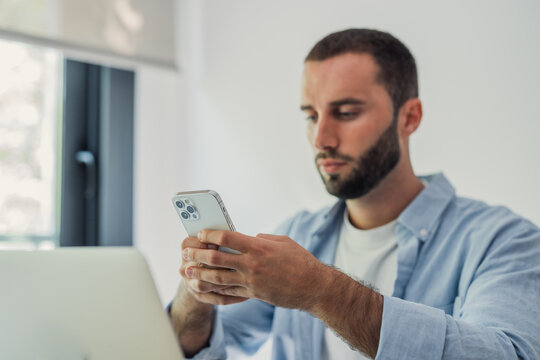 Young man using a smartphone while sitting in front of a laptop, engaging with multiple digital devices for communication, work, or social media in a modern setting