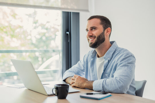 Young bearded man smiling and looking away from the camera, sitting at a wooden desk with a laptop, cup, and notebook, enjoying a productive remote work-from-home setup