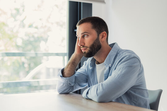 Young handsome man with beard looking away through a bright window, resting his head on his hand while contemplating, feeling worried, lonely, or thoughtful indoors