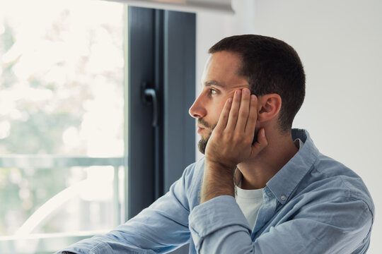 Young man thinking and looking away through a window. Resting his face on his hand. Reflecting on thoughts while being alone in an indoor office or home setting. Experiencing anxiety. Solitude