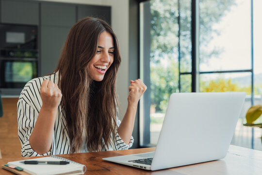 Woman feeling excited and happy, raising fists in a gesture of triumph while looking at a laptop screen, achieving a goal, or winning an online competition through her remote work setup