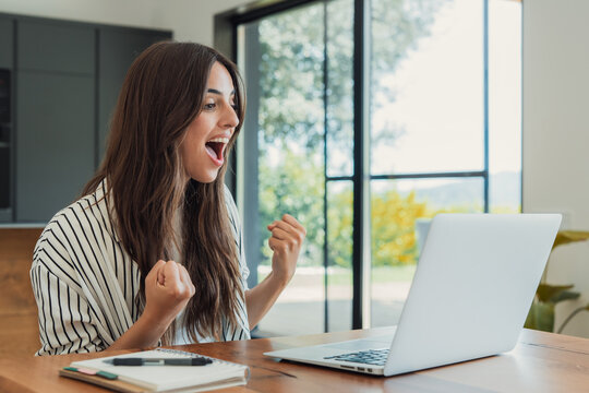 Woman feeling excited and celebrating success while looking at a laptop computer, working remotely from her modern home office during the day