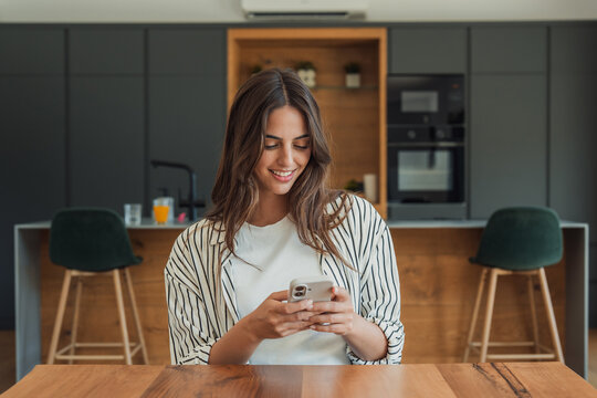 Young woman sitting at a table in a stylish kitchen, happily interacting with her mobile device, representing connectivity, communication, and modern living