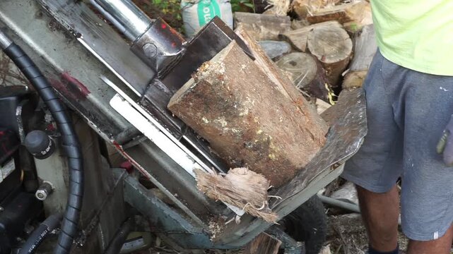 Close-up view of a worker using a powerful hydraulic log splitter machine to split a large wooden log into smaller firewood pieces outdoors.