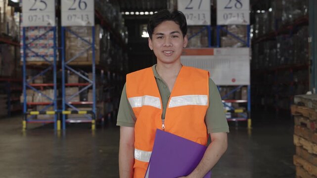 Smiling Asian man presents purple folder within brightly lit warehouse environment showcasing organized pallet storage and labeled shelving systems for industrial operations support.