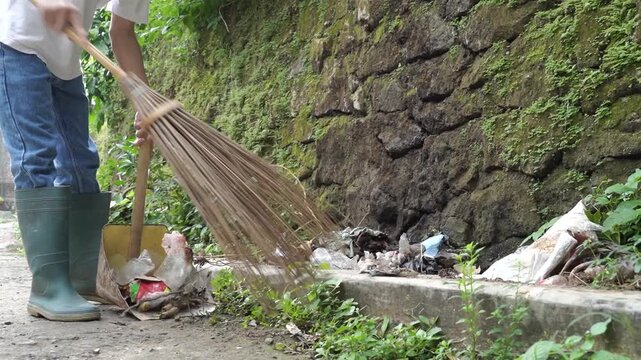 A person cleans the outdoor street scene, sweeping along a curb beside a rough stone wall