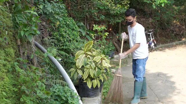 A man cleans a garden path outdoors with a broom during daylight. The main action shows sweeping along a narrow concrete walkway bordered by lush greenery