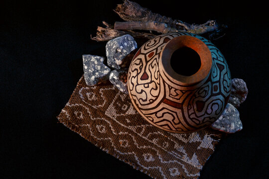 A traditional clay vessel from the Peruvian jungle, featuring intricate, undulating designs in shades of brown and turquoise. Rough stones and dried branches surround it on a dark fabric. The geometri