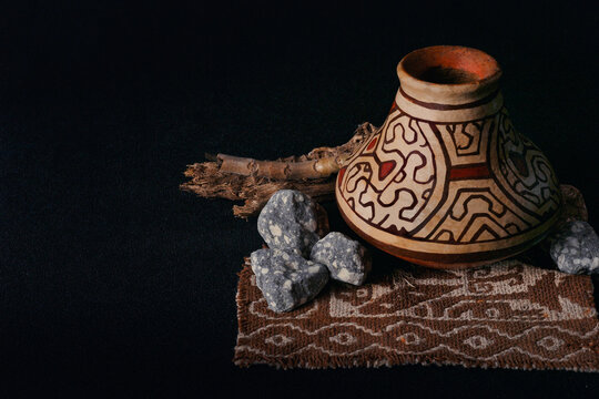 Ancient clay pot gleams with intricate designs. Rough stones and twig rest beside it. Patterned textile anchors the composition. Deep black background enhances texture and detail