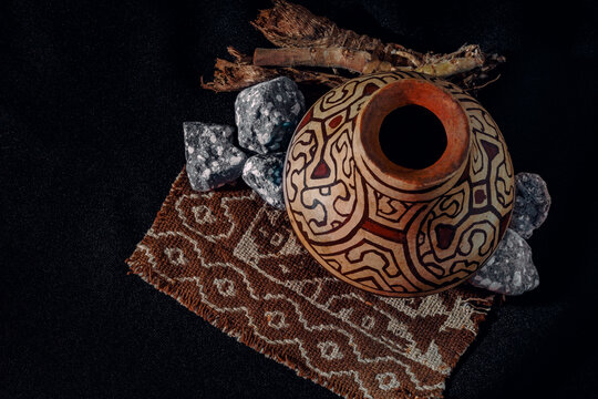 Traditional clay vessel from the Peruvian jungle, with intricate spiral patterns. The dark background highlights the pieces. Ancient stones and woven fabric frame the vessel.