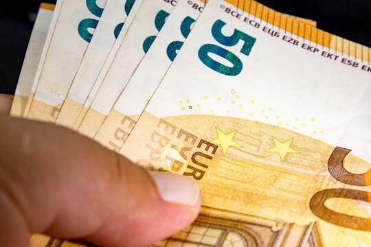 Fingers grasping several euro currency notes, showcasing detailed design elements including stars, text, and architectural features. The euro banknotes display vibrant colors and security markings, em