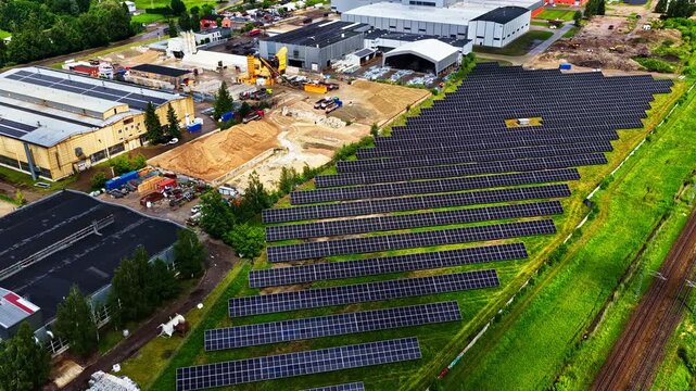 Solar panel field beside industrial buildings generating renewable energy preparatory for new build construction demand, aerial static as cloud shadows pass