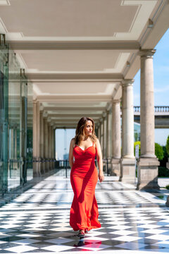 Young smiling latina woman walking confidently and gracefully relaxed posture through a bright corridor at Castillo de Chapultepec in Mexico City, wearing a fitted vibrant red dress with elegant style
