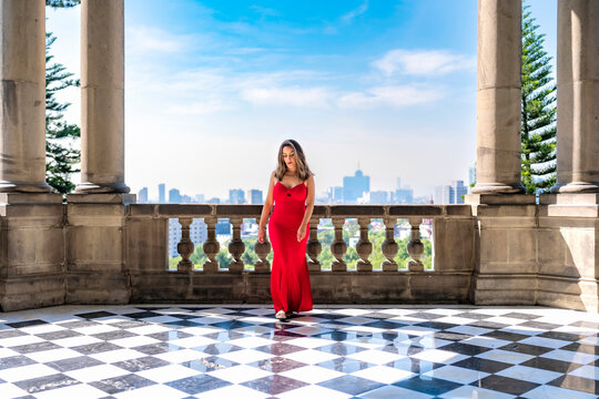 Young smiling latina woman posing confidently and calmly, enjoying the view while standing on a balcony at Castillo de Chapultepec in Mexico City, wearing an elegant long red dress