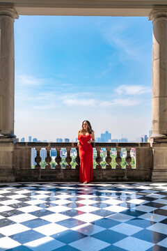 Young smiling latina woman posing confidently and calmly, enjoying the view while standing on a balcony at Castillo de Chapultepec in Mexico City, wearing an elegant long red dress