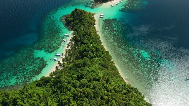 Overhead flyover above forested Maxima Island toward Cayoya Island with white sand spit outrigger boats and turquoise reef in Philippines