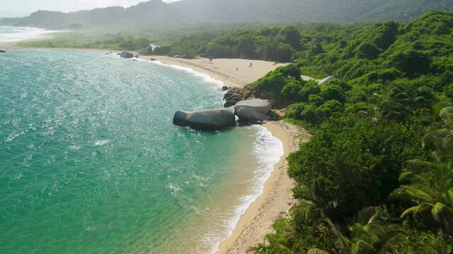 Aerial flyover of Playa Arenilla curved sandy beach with giant granite boulders turquoise water and jungle mountains at Parque Tayrona near Santa Marta Colombia