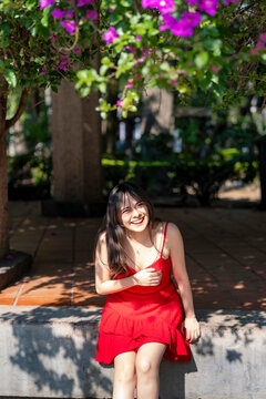 Young smiling latina woman sitting relaxed, enjoying soft sunlight and shadows at Parque Mexico in Condesa, Ciudad de Mexico surrounded by colorful flowers, wearing a red dress, delicate accessories