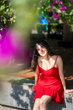 Young smiling latina woman sitting relaxed, enjoying soft sunlight and shadows at Parque Mexico in Condesa, Ciudad de Mexico surrounded by colorful flowers, wearing a red dress, delicate accessories