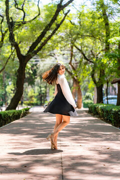 Young smiling latina woman twirling playfully, cheerful and carefree outdoors at Parque Mexico in Condesa, Ciudad de Mexico surrounded by trees sunlight, wearing a black dress, white blouse in motion