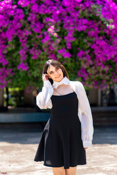 Young smiling latina woman posing relaxed, cheerful outdoors at Parque Mexico in Condesa, Ciudad de Mexico surrounded by vibrant bougainvillea flowers, wearing a black dress, white blouse, sunlight