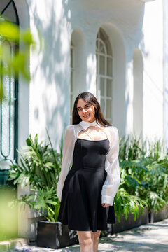 Young smiling latina woman posing relaxed, cheerful on streets near Parque Mexico in Condesa, Ciudad de Mexico surrounded by white architecture greenery, wearing a black dress, white blouse, sunlight