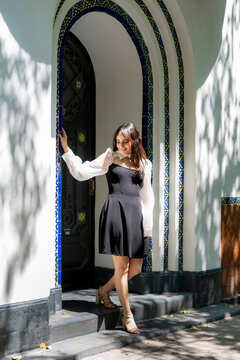 Young smiling latina woman posing relaxed, cheerful on streets near Parque Mexico in Condesa, Ciudad de Mexico surrounded by white architecture greenery, wearing a black dress, white blouse, sunlight