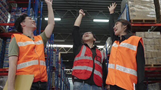 Three employees collaborate within brightly lit warehouse amidst stacked pallets and boxes, showcasing teamwork and positive industrial operations wearing high-visibility vests.