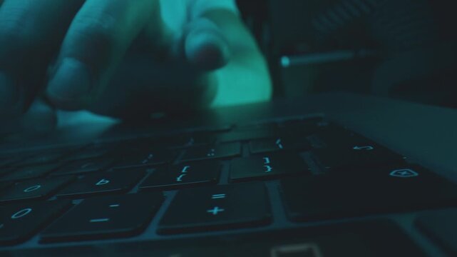 Close up of male hands typing on laptop keyboard in dark room with blue lighting, focused on digital work.