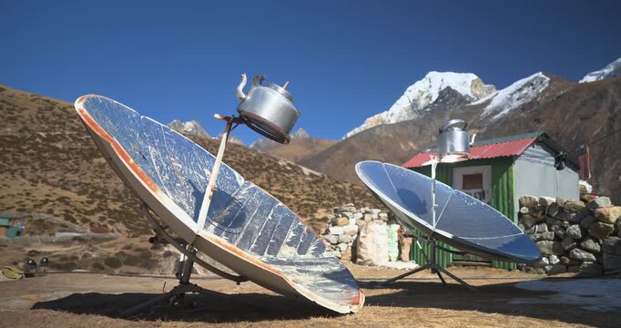 A static wide-angle shot of reflective parabolic solar cookers boiling water in kettles at a mountain village teahouse lodge, with Ama Dablam peak visible in Namche Bazaar, Everest Region of Nepal