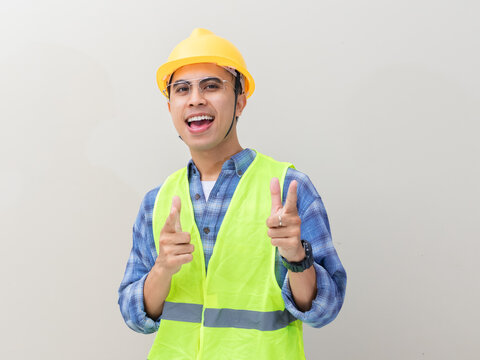 Excited young Asian engineer wearing a yellow hard hat and safety vest pointing forward to invite viewers against a plain background