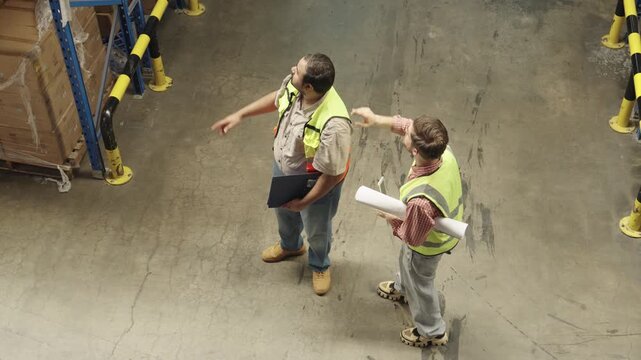 Two professionals discuss warehouse blueprints and safety procedures within dark industrial facility, one holding technical drawings while the other wears safety vest and clipboard.