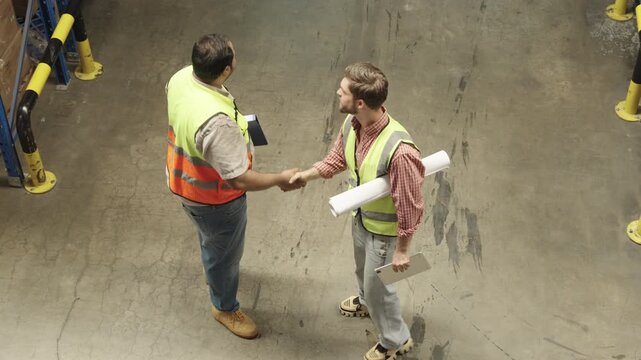 Two men wearing yellow vests discuss blueprints and industrial equipment within spacious warehouse setting, showcasing engineering processes and inspection activities alongside shelving units.