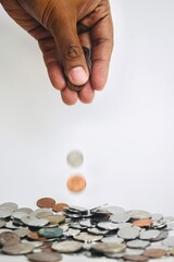 A hand releasing two coins captured mid-air as they fall toward a large pile of scattered Malaysian ringgit coins on a white surface, illustrating rapid growth, micro-investments, or tipping. © Akram