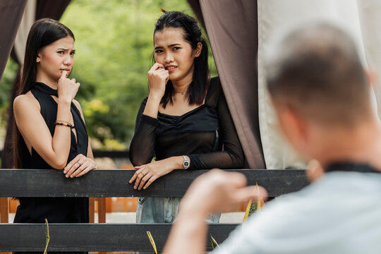 Two young Southeast Asian women react with awkward and uncomfortable expressions while an older man waves at them outdoors.