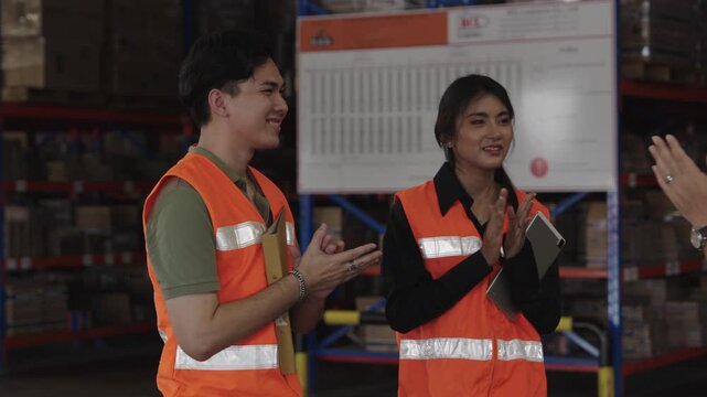 Two workers inspect documents and folder within dark warehouse filled with stacked pallets; they wear orange safety vests and reflective gear for industrial inspection purposes.