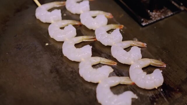 High-angle close-up of a row of fresh, raw, peeled and deveined white shrimp being cooked on a hot metal flat-top grill. The shrimp are translucent and ready to be seared as part of a professional cul
