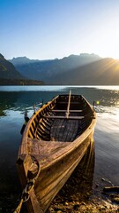 Fototapeta premium Old wooden rowboat moored on a calm lake at sunrise with mountains in the background.